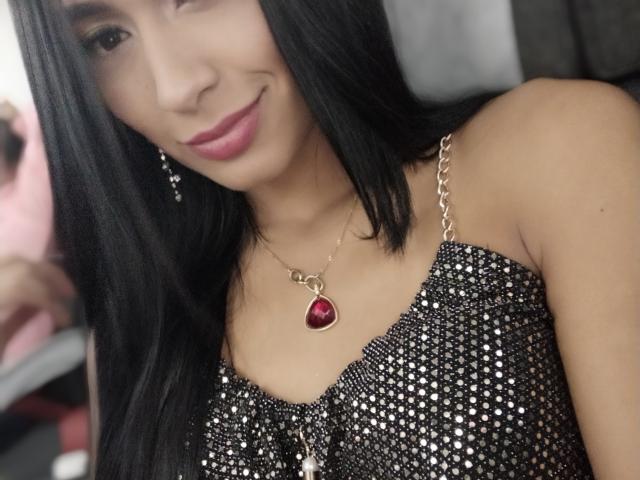 ValeriaKlaus with black hair, pink lipstick, wearing a sparkly black dress and a red heart necklace, smiling at the camera, showing her tits slightly visible under the dress.