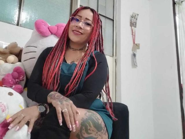 LaurenBlake with red braided hair, glasses, black cardigan, teal dress, tattoos on hands and thigh, sitting on chair, smiling, pink Hello Kitty plush in background.