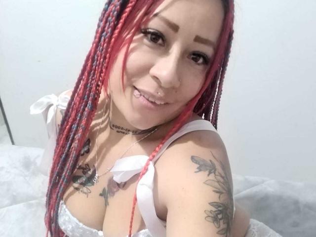 LaurenBlake with red and black braids, nose ring, and tattoos, smiling, showing her tits in a white lace top. She's on a bed with a gray blanket.