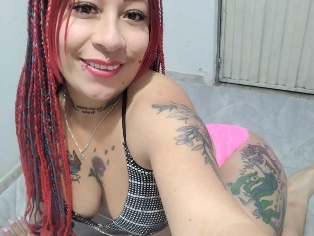 LaurenBlake with red braids, nose ring, and pink panties, smiling, showing her tattoos, tits, and ass, kneeling on gray tiled floor.