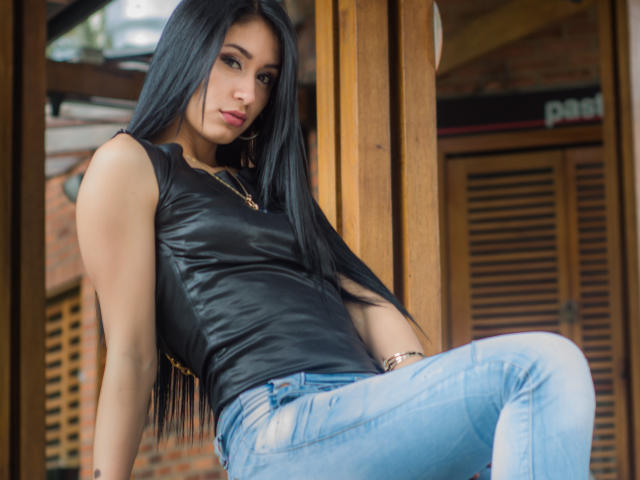 ValeriaKlaus with long black hair, wearing a black leather sleeveless top and blue jeans, leaning against wooden beams, looking seductively at the camera. Her ass is slightly raised.