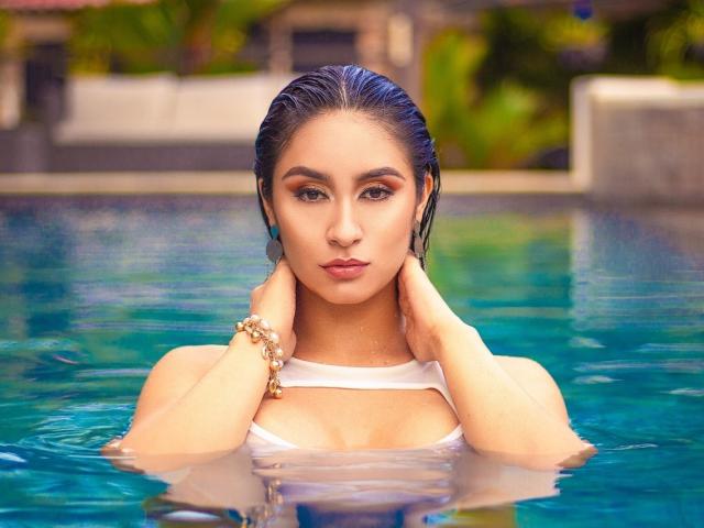 ValeriaKlaus with wet, slicked-back dark hair, wearing a white top, hands on her neck, in a blue pool, looking at the camera with a serious expression, showing her tits.