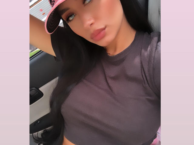 ValeriaKlaus with long black hair, wearing a pink cap, grey shirt, showing her big tits, pouty lips, and blue eyes, in a car, looking at the camera.