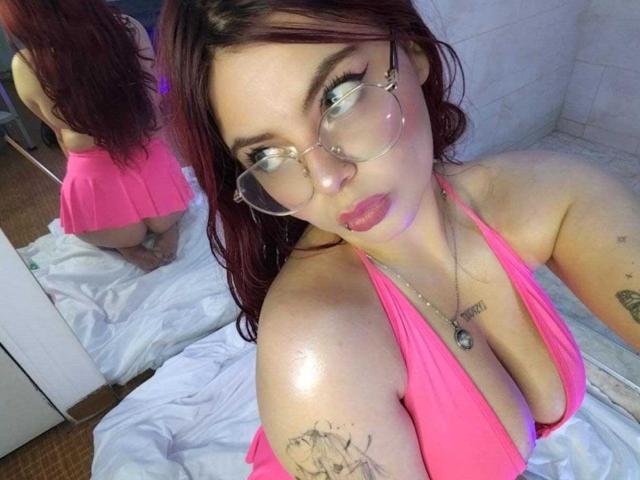MelanieGate with red hair, glasses, and pink halter top showing big tits, sitting on bed, reflection in mirror shows her ass, tattoos on arm, looking up with pouty lips.