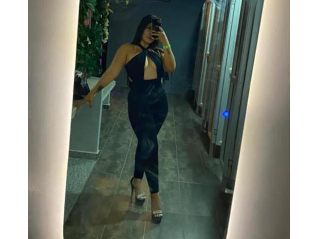 Camila Verone, vestida con un conjunto negro, con aberturas, de cintura alta y ajustado, de pie y con tacones, tomando una foto de sí misma en el espejo, mostrando sus curvas, su trasero tonificado y sus pequeños senos..