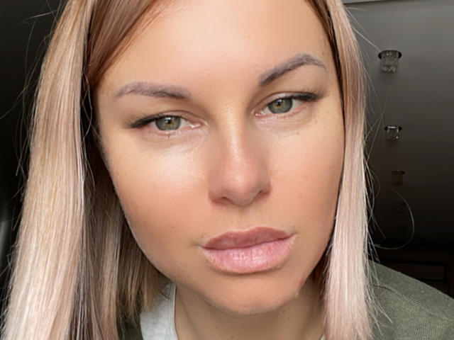 El rostro de BelleLisaG, con labios rosados y ligeramente prominentes, ojos verde claro y cabello rubio, tiene una expresión ligeramente molesta o seria, y lleva una camisa verde en un primer plano..