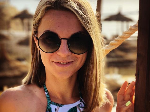 BelleLisaG con cabello rubio, usando gafas de sol redondas y oscuras, sonriendo, sosteniendo una cuerda, con un top de tirantes de colores, mostrando ligeramente sus pechos a la luz del sol..