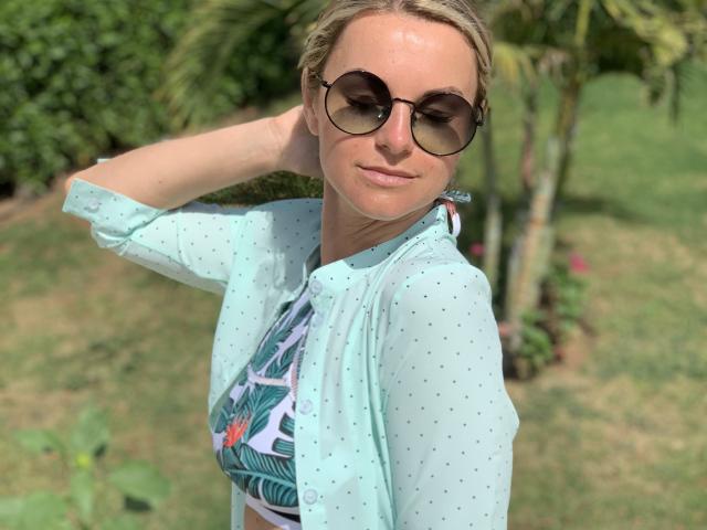 BelleLisaG com óculos de sol, camisa azul com bolinhas, e top de biquíni com estampa tropical, mão no cabelo, sorrindo, com luz solar, fundo de jardim verde..