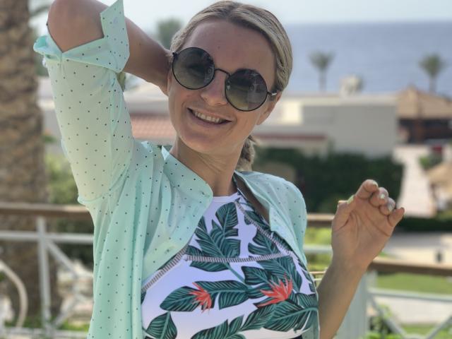 BelleLisaG met blond haar in een vlecht, die een groene stippen blouse draagt over een top met een tropisch bladprint, en donkere ronde zonnebril, lachend met één hand in haar haar..