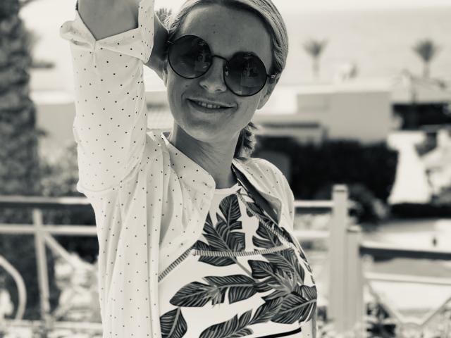BelleLisaG with sunglasses, smiling, wearing a patterned shirt and leafy top, raising arm, holding hair back, showing medium tits, black-and-white photo, casual, outdoor setting.