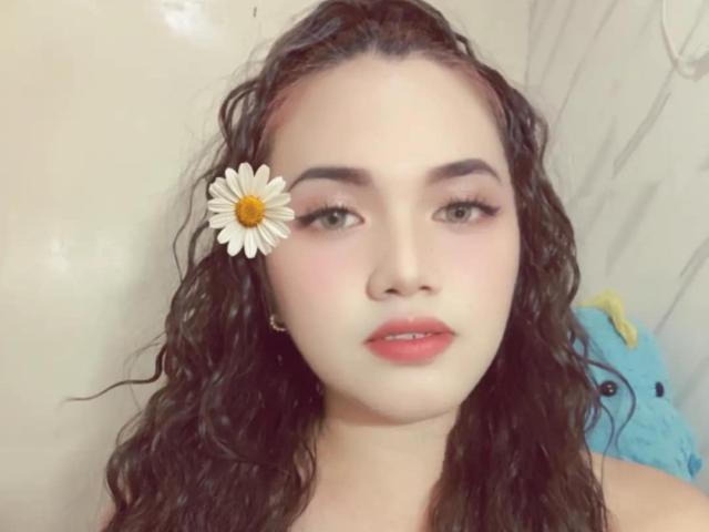 AsianHotCock69 has wavy black hair, a white daisy in her hair, pink lipstick, and green eyes. She's looking at the camera with a soft, slightly pink face.