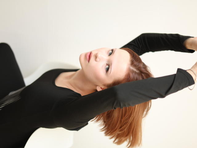 MollyWettmond has red hair, is wearing a black long-sleeve top, and is stretching with arms raised, looking at the camera with a slight smile.