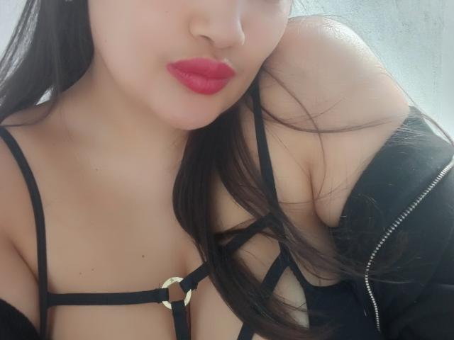 ZafiRouge con lápiz labial rojo, un sujetador negro con tirantes que deja ver sus pechos, una chaqueta negra llevada fuera del hombro, cabello largo y oscuro, piel clara, luz suave, primer plano, sexy, labios ligeramente fruncidos, expresión suave..