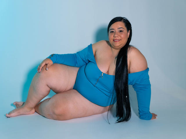 AndreaaX69, curvy with large tits and ass, sits on floor in blue off-shoulder top, long black braid, smiling, legs bent, right arm resting, left arm on knee.