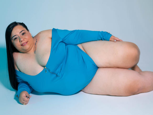 AndreaaX69, de talla grande, con busto generoso y muslos gruesos, acostada en un vestido azul con un hombro descubierto, sonriendo a la cámara, cabello negro largo, tatuaje en el hombro derecho..