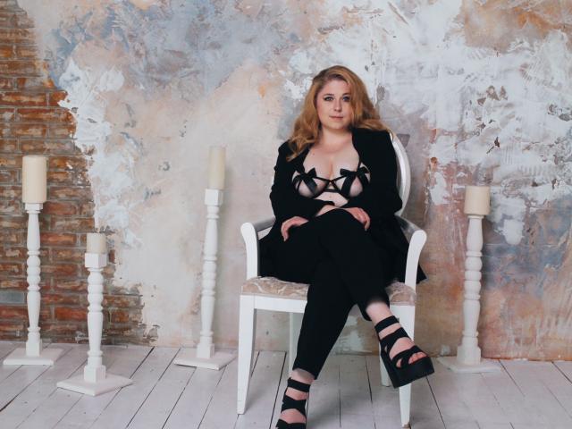 JessikaDaniels, blonde, assise sur une chaise blanche, portant un haut noir à bretelles laissant entrevoir ses seins, un pantalon noir et des talons compensés noirs, avec une expression sérieuse, entourée de bougies..