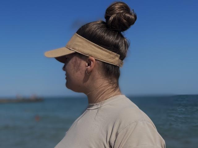 Madrinia with a bun, wearing a beige visor and light shirt, has her ass slightly visible, standing by the ocean under a clear blue sky.