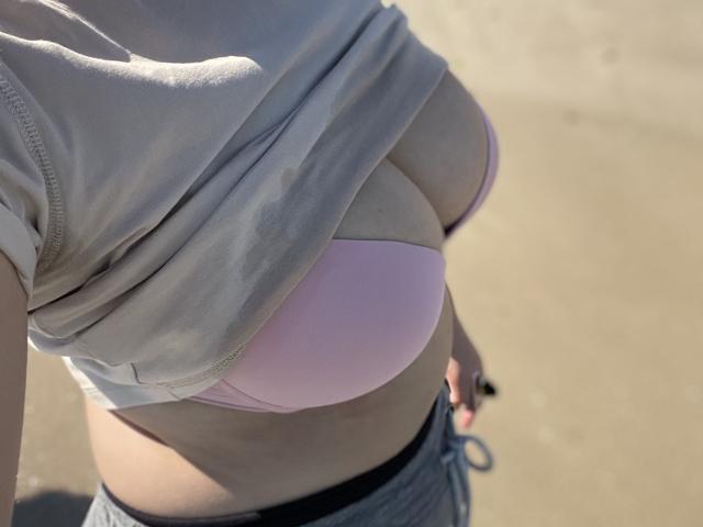 Madrinia's large tits in a pink bra, covered by a lifted gray shirt, showing pale skin and gray sweatpants with a black waistband. Sunlight casts shadows.