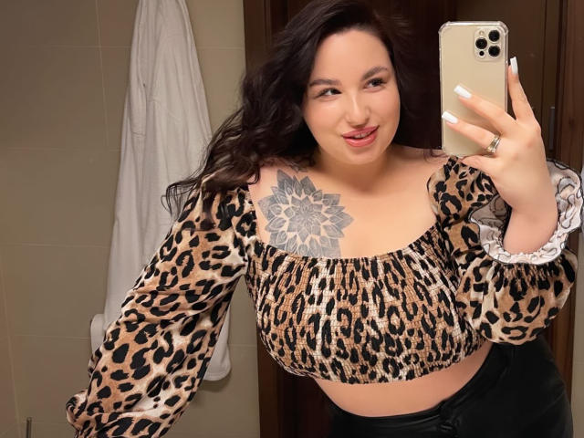 CuteBeatrice trägt ein Oberteil mit Leopardenmuster, das ihre großen Brüste zeigt, und hat ein Blumen-Tattoo an ihrer rechten Schulter. Sie macht ein Selfie vor einem Spiegel mit einem verschmitzten Lächeln und hält ein goldenes iPhone..