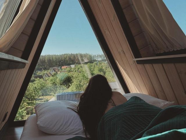 A CuteBeatrice, com seus longos cabelos castanhos, está deitada em uma cama, virada para uma janela com árvores e casas verdes do lado de fora, coberta com um cobertor verde, sob cortinas brancas..