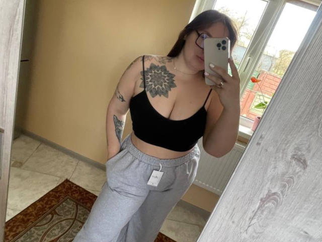 CuteBeatrice con il seno prosperoso, indossa una crop top nera, pantaloni da jogging grigi, ha dei tatuaggi, occhiali e tiene un telefono, sta scattando un selfie allo specchio, mostrando la mano in tasca..