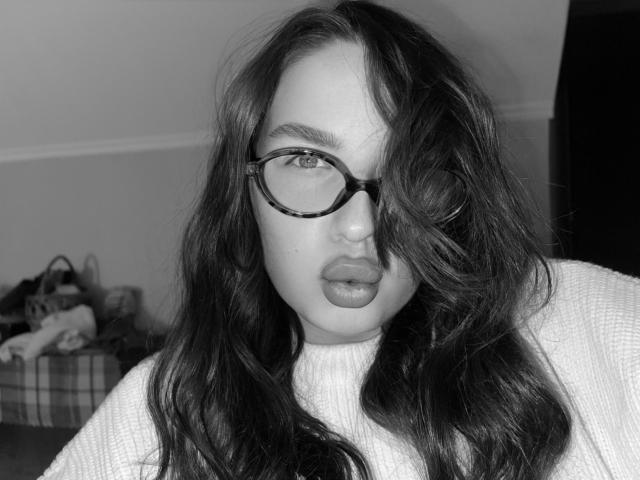 CuteBeatrice tiene el pelo oscuro y ondulado, gafas de montura de carey y labios carnosos. Lleva un jersey grueso de punto, y sus ojos están ligeramente ocultos por su cabello. Foto en blanco y negro..