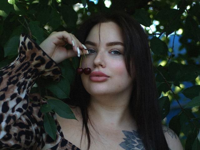 CuteBeatrice, con lunghi capelli castani, occhi azzurri e una maglietta a stampa leopardo, tiene una ciliegia vicino alla sua bocca leggermente aperta e colorata di rossetto rosa. Ha un tatuaggio sul petto..