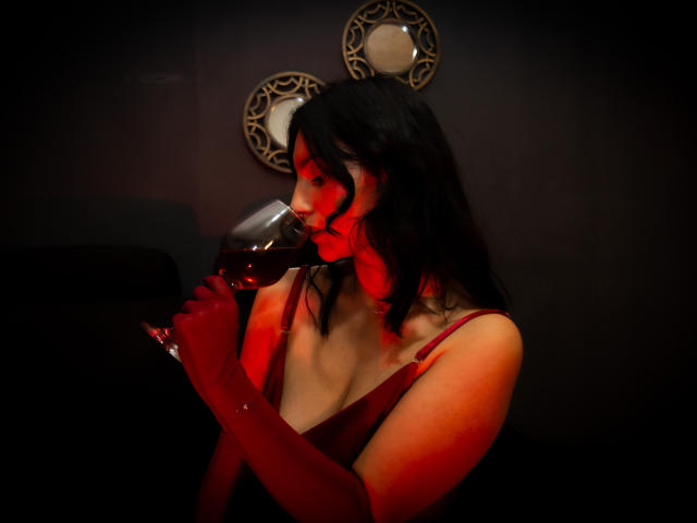Sammy Hoffman, con i capelli neri ondulati, un vestito rosso e guanti rossi, sorseggia un vino rosso scuro, illuminata da una luce rossa che le dona un bagliore, tiene il bicchiere alle labbra, con il seno visibile..