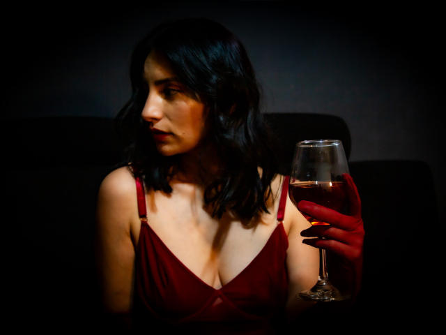 Sammy Hoffman, avec ses cheveux noirs et ondulés, sa robe rouge et ses gants rouges, tenant un verre de rosé, regardant sur le côté, dans une lumière tamisée, montrant légèrement sa poitrine..