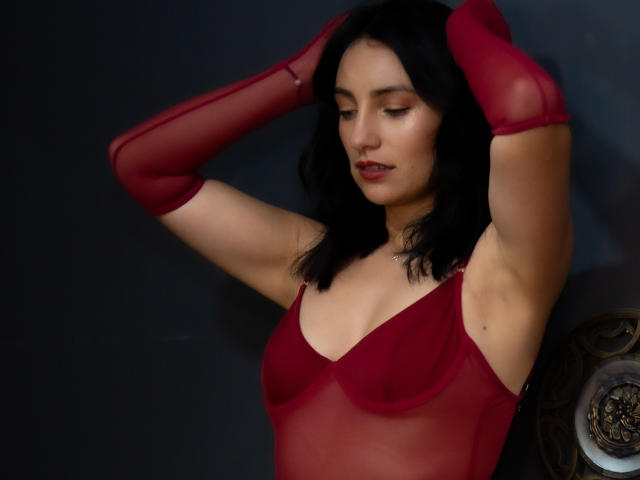 Sammy Hoffman, com cabelo escuro, uma blusa vermelha transparente e mangas que combinam, braços levantados, mostrando os seios, e uma expressão sutil e confiante. Ela está em frente a um fundo escuro..