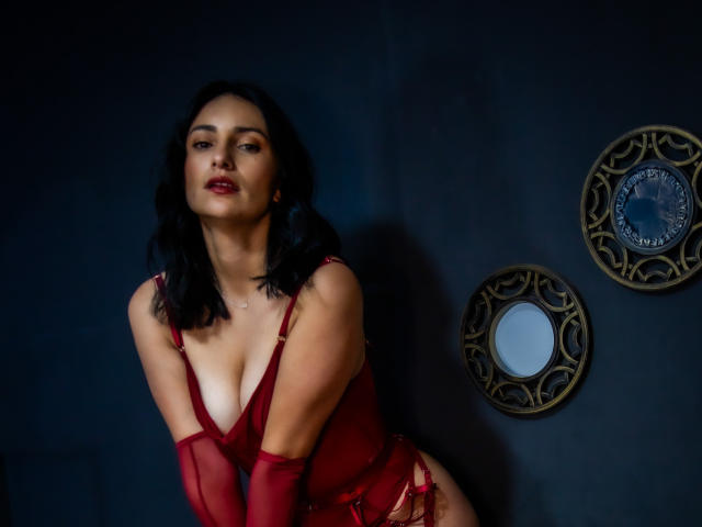 SammyHoffman with black hair, red lingerie, and gloves, leaning forward, showing her tits and red-painted lips, in dim lighting, with two ornate mirrors on a dark wall.