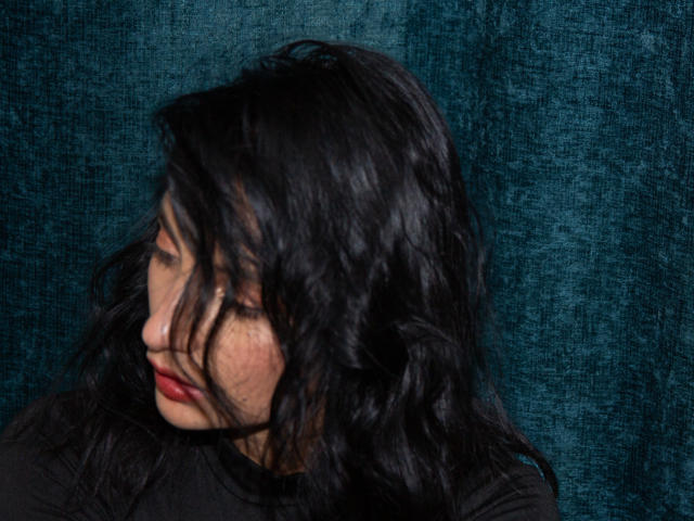 SammyHoffman's wavy black hair covers her eyes, red lips slightly parted, looking down with a subtle smile, against a dark teal textured background. She's wearing a black top.