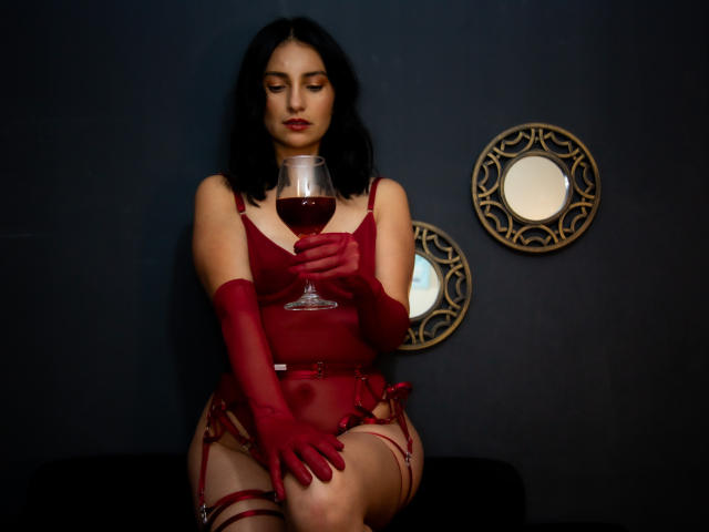 SammyHoffman with dark hair, red lingerie, garters, and gloves, holding a glass of red wine, sitting, looking down, in a dark room with circular mirrors.