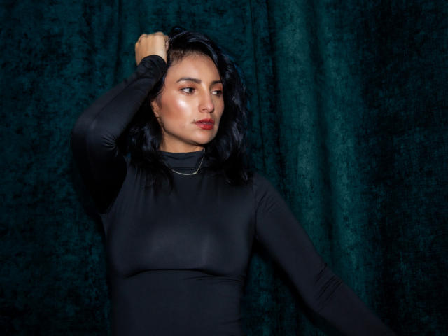SammyHoffman with dark wavy hair, wearing a tight black long-sleeve shirt, looking to the side, hand in hair, green velvet background, subtle makeup, red lips.