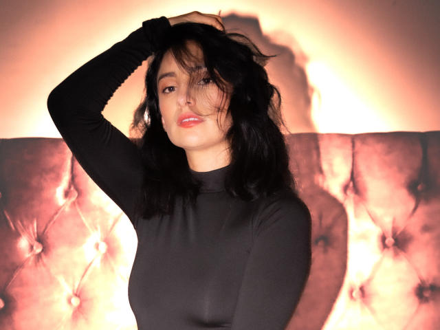 Sammy Hoffman, avec des cheveux noirs, portant un haut noir à manches longues, une main dans les cheveux, regardant la caméra, lumière orange douce, tête de lit capitonnée, ombre sur le mur. Seins visibles sous le haut..