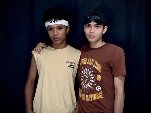 GeroandMax met krullend zwart haar, donkere huid, een witte hoofdband, een beige shirt, met een arm om de bruingehuidige Max in een bruine pet en een shirt met een zonprint, staand voor een donkere achtergrond..