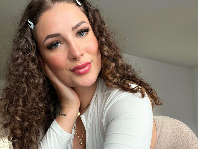 KateCurl-hot with curly brown hair, white hair clips, pink lipstick, and a white top, resting her cheek on her hand, smiling at the camera, showing her cute ass.