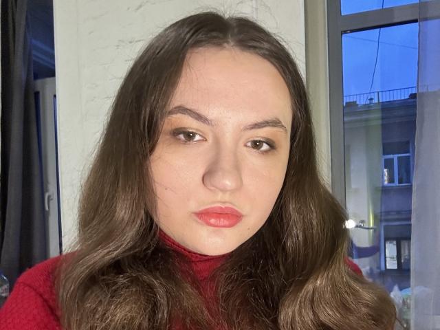LuluKatrina has long brown wavy hair, red lips, and brown eyes. She's wearing a red turtleneck, looking serious, with a slightly pouty expression. Her tits are not visible.