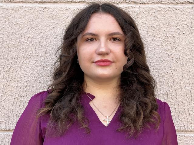 LuluKatrina has wavy brown hair, pink lips, a small mole on her right cheek, and wears a purple shirt with a cross necklace, standing in front of a textured wall.