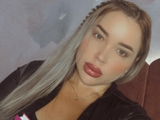 MiaBlondeHot with pouty red lips, long blonde braided hair, brown eyes, and a black top, showing her chest and necklace. Her tits are partially visible.
