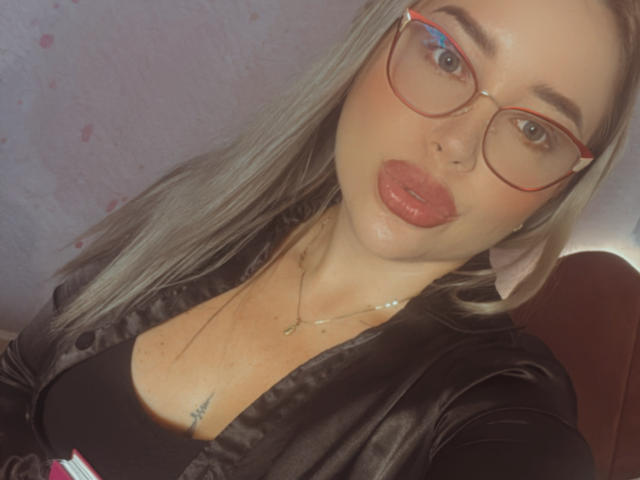 MiaBlondeHot with puffy red lips, wearing red glasses, black satin robe, black top, and necklace, showing a small tattoo on her chest. Blonde hair, fair skin, cute.