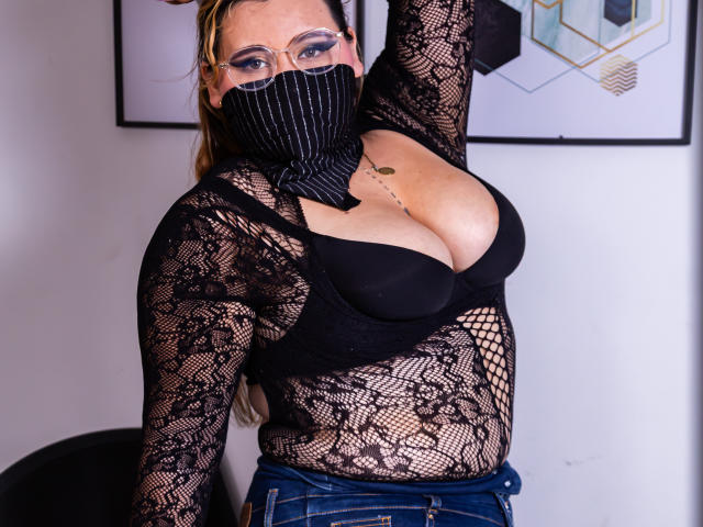 SalmaAzaman with big tits in black lace top, fishnet sleeves, and pinstriped mask, arm raised, wearing blue jeans, glasses, and gold necklace, standing in front of wall.