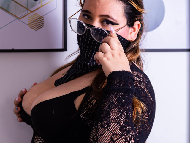 SalmaAzaman with glasses, pinstripe mask, black lace top, showing big tits, holding mask with right hand, brown hair, white wall, framed art in background.