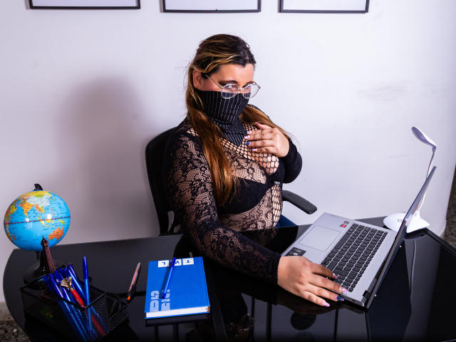 SalmaAzaman, wearing a black lace long-sleeve top, glasses, and mask, touches her chest while typing on a laptop, with a globe and pens on a black desk.
