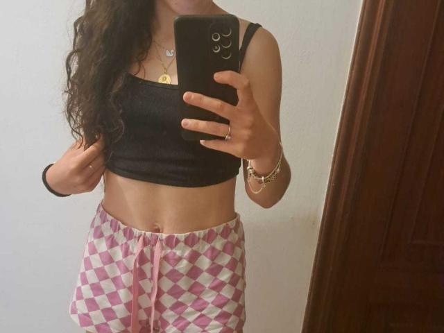 ParejaCachonda69 with long curly brown hair, wearing a black tank top, pink and white diamond-patterned pants, taking a mirror selfie, showing a slim, toned midriff.