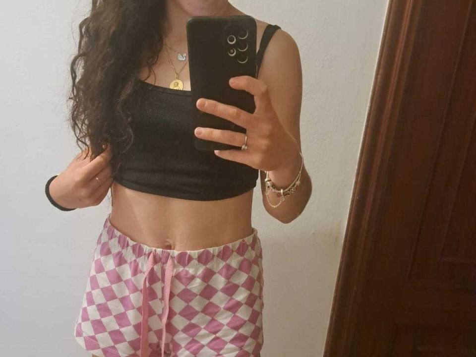 ParejaCachonda69 with long curly brown hair, wearing a black tank top, pink and white diamond-patterned pants, taking a mirror selfie, showing a slim, toned midriff.