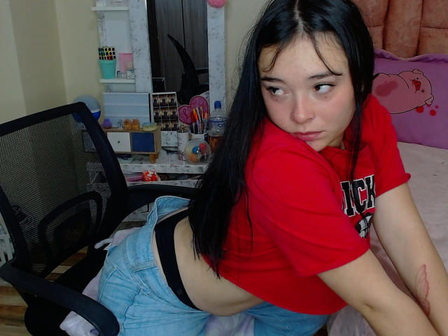 Jhanan, con cabello negro, una camisa roja y jeans azules, apoyada en una cama, mostrando su ropa interior negra y un pequeño tatuaje en su brazo, con una mirada sensual y de lado..