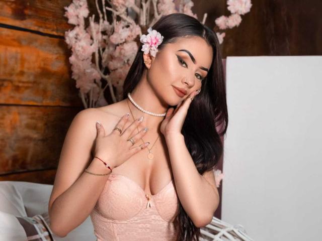 KattRouse ha lunghi capelli scuri, un fiore rosa tra i capelli, e indossa un reggiseno rosa in pizzo, una collana di perle e degli anelli. Sta toccando la guancia con la mano destra..