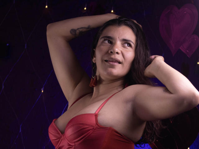 BulmaHot with medium tits, red dress, arms raised, tattoo on left arm, smiling, brown hair, dark eyes, red earrings, purple background, heart shapes, slightly chubby, cute.