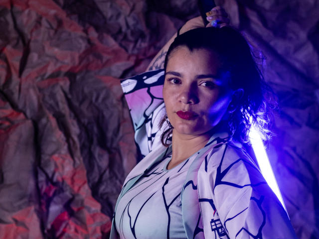 BulmaHot with curly hair, wearing a white kimono with black lines, holding a glowing blue sword, serious expression, red lipstick, rocky background, medium shot, slightly illuminated from side.